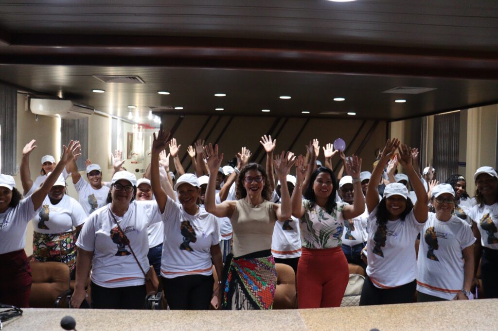 Em sala fechada vê-se grupo de mulheres com camisas brancas do Projeto Semente Crioula. Ao centro encontra-se Livia Viana e Susan Lucena. Todas as presentes estão com os braços levantados e sorrindo para a câmera.