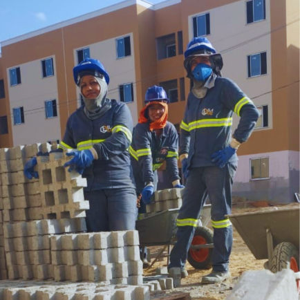 Três mulheres em canteiro de obra usando capacete, luvas e uniforme atrás de bloquetes de concreto