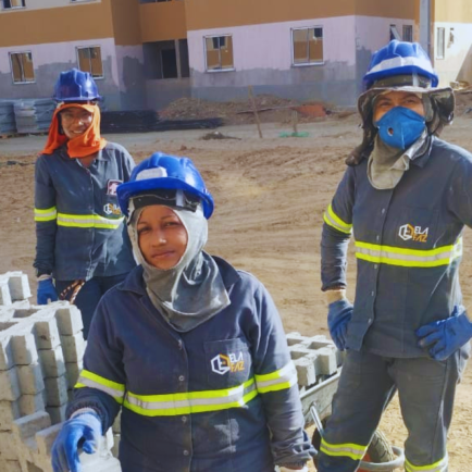 Três mulheres em canteiro de obra usando capacete, luvas e uniforme ao lado de bloquetes de concreto